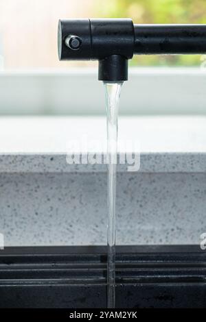 Wasser fließt aus einem schwarzen Küchenhahn. Der Wasserhahn befindet sich vor einem Küchenfenster über einem gesprenkelten Quarzsprenkel Stockfoto