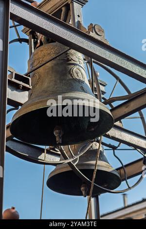 Zwei große Bronzeglöckchen mit Klappern hängen in einem Metallgürtel und warten darauf, geläutet zu werden. Die Glocken sind mit aufwändigen Schnitzereien und lateinischer Inschrift verziert Stockfoto