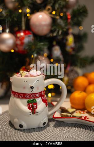 Nahaufnahme eines niedlichen Eisbärenbechers, der vor einem verzierten Weihnachtsbaum mit festlichen Ornamenten steht. Konzept von gemütlichen Winterferien und warmen Getränken Stockfoto
