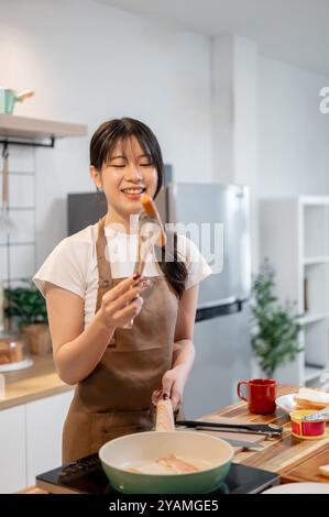 Eine schöne, glückliche asiatische Frau in einer Schürze genießt es, in der Küche Frühstück zu kochen und Würstchen in einer Pfanne zu braten. Hausmannskost, Hausleben, Leute Stockfoto