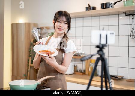 Eine hübsche und glückliche junge asiatische Essenskünstlerin oder Chefkoch in einer Schürze spricht und nimmt ein Video auf, während sie in der Küche kocht und zeigt Stockfoto
