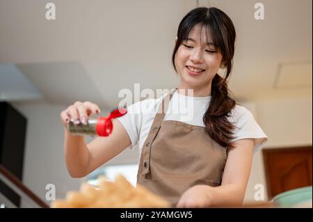 Eine schöne, glückliche junge asiatische Frau in einer Schürze streut Pfeffer auf ihr Essen und genießt es, in der Küche zu Hause zu kochen, während sie das Frühstück zubereitet. Stockfoto