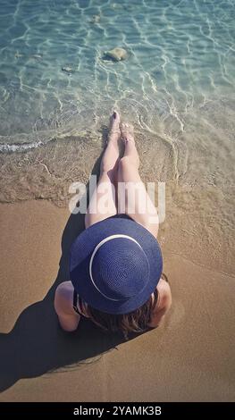 Rückansicht junge Frau mit blauem Sonnenhut am Strand mit gestreckten Beinen zum Meer hin. Das klare Wasser schlägt sanft mit ihren Füßen. Sere Stockfoto