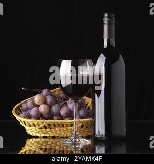 Eine volle Flasche Rotwein, ein Glas Wein und schwarze Trauben in einem Korb auf schwarzem Hintergrund Stockfoto