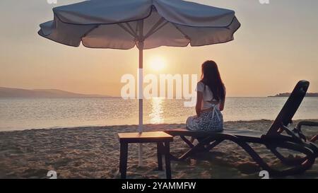 Ästhetische junge Frau, die auf einer Sonnenliege am Strand saß und die Morgensonne mit Blick auf das Meer beobachtete. Wunderschöne Szenerie am Meer, summ Stockfoto