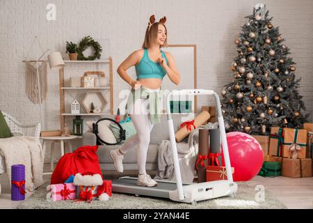 Sportliche junge Frau mit Hirschgeweih und Weihnachtsgeschenken, die zu Hause auf dem Laufband laufen Stockfoto