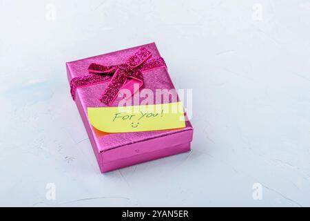 Eine leuchtend pinke Geschenkbox, die mit einem glänzenden Band und einer Notiz gebunden ist, auf einem sanften hellen Hintergrund platziert und symbolisiert Feiern und Geschenke. Stockfoto