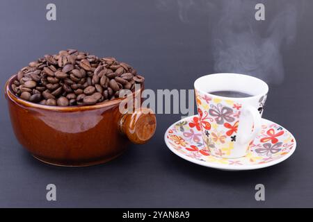 Eine mit frisch gerösteten Kaffeebohnen gefüllte, warme braune Schüssel gibt neben einer dampfenden Tasse gebrühten Kaffee vor schwarzem Hintergrund Dampf ab Stockfoto