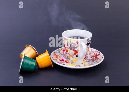 Neben Kaffeekapseln steht eine dampfende Tasse frisch gebrühten Kaffees und schafft eine einladende Atmosphäre vor einem eleganten schwarzen Hintergrund Stockfoto