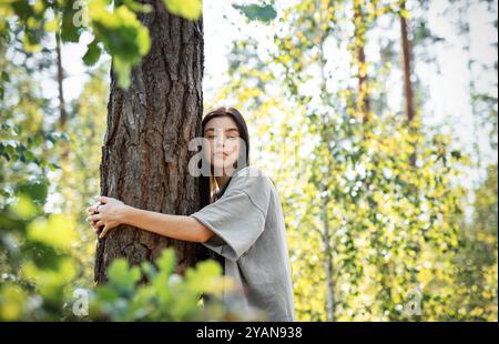 Eine Frau lehnt sich an einen Baum, blickt mit einem ruhigen Ausdruck nach oben, umgeben von leuchtend grünen Blättern in einem sonnendurchfluteten Wald. Stockfoto