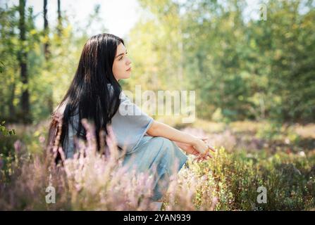 Eine junge Frau sitzt ruhig auf dem Waldboden, ihr langes Haar zieht sich über den Rücken. Sie erscheint tief in Gedanken, umgeben von blühenden Wildblumen Stockfoto