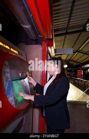 Junge Frau in Geschäftskleidung, die nachts am Bahnhof am Fahrkartenautomaten vorbeifährt Stockfoto