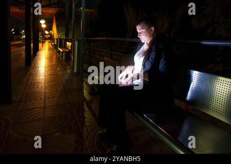 Oung Frau in Geschäftskleidung, die am Laptop arbeitet, während sie am Nachtbahnhof auf den Zug wartet Stockfoto