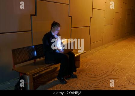 Oung Frau in Geschäftskleidung, die am Laptop arbeitet, während sie am Nachtbahnhof auf den Zug wartet Stockfoto