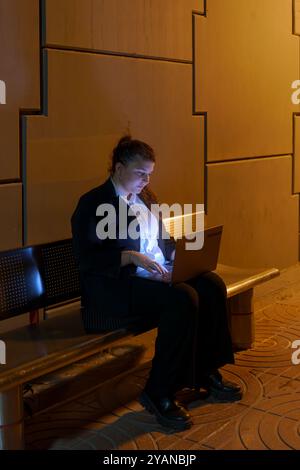 Oung Frau in Geschäftskleidung, die am Laptop arbeitet, während sie am Nachtbahnhof auf den Zug wartet Stockfoto