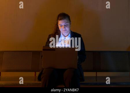 Oung Frau in Geschäftskleidung, die am Laptop arbeitet, während sie am Nachtbahnhof auf den Zug wartet Stockfoto