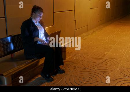 Oung Frau in Geschäftskleidung, die am Laptop arbeitet, während sie am Nachtbahnhof auf den Zug wartet Stockfoto