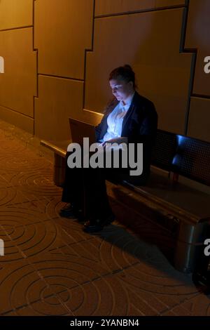 Oung Frau in Geschäftskleidung, die am Laptop arbeitet, während sie am Nachtbahnhof auf den Zug wartet Stockfoto