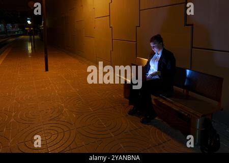 Oung Frau in Geschäftskleidung, die am Laptop arbeitet, während sie am Nachtbahnhof auf den Zug wartet Stockfoto