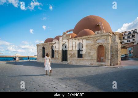 Eine Frau allein reist, Rückansicht einer jungen Frau in weißem Sommerkleid, die an der Kucuk-Hasan-Pascha-Moschee im Hafen von Chania, Kreta, Griechenland, vorbeiläuft Stockfoto