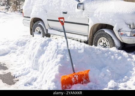 An einem sonnigen Wintertag ragt eine orange Schneeschaufel vor einem schneebedeckten weißen SUV-Auto aus einer Schneewolke hervor Stockfoto