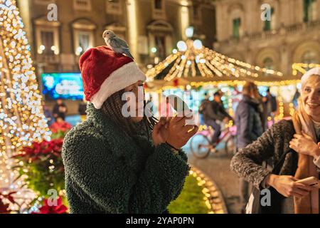 Eine junge Frau mit Weihnachtsmütze auf dem Weihnachtsmarkt mit einem Vogel auf dem Kopf. Festliche Atmosphäre mit Weihnachtslichtern, Dekorationen und Menschen, die das genießen Stockfoto