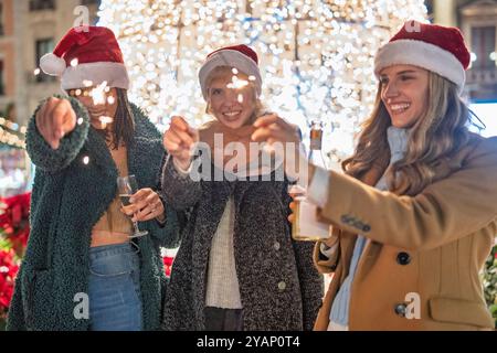 Verschiedene Gruppen junger Frauen feiern Weihnachten auf einem Festmarkt. Freunde im Weihnachtsmann lächeln und toasten mit Getränken umgeben von Feiertagslicht Stockfoto