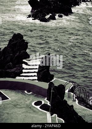 Schwarzweiß-Aufnahme von Porto Moniz, wo der wilde Ozean auf künstliche Treppen und Tauchbretter an Madeiras rauer Küste trifft Stockfoto