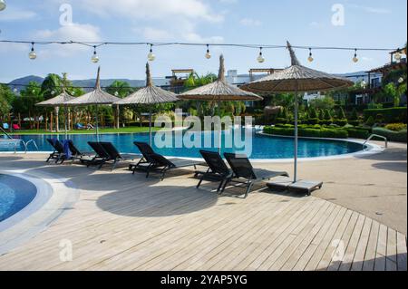 Der ruhige Poolbereich bietet komfortable Liegestühle und bezaubernde Strohschirme, die zum Entspannen unter klarem, blauem Himmel und sanftem Sonnenlicht einladen Stockfoto