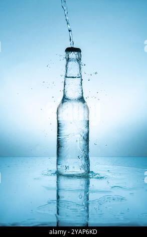 Wasserstrahl spritzt auf Glasflasche mit mit Wasser gefülltem Verschluss auf feuchter reflektierender blauer Oberfläche vor blauem Hintergrund Stockfoto