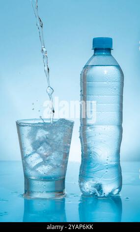 Wasserstrahl spritzt auf gefrorenen Glasbecher, gefüllt mit Wasser und Eis, neben Plastikflasche auf feuchter reflektierender blauer Oberfläche vor blauem Hintergrund Stockfoto