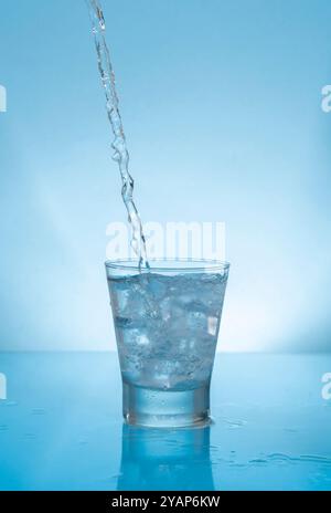 Wasserstrahl spritzt auf Glasbecher gefüllt mit Wasser und Eis auf feuchter reflektierender blauer Oberfläche vor blauem Hintergrund Stockfoto