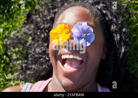 Verspielte positive afroamerikanische Frau bedeckt die Augen mit zwei Blumen, genießt Frühlingszeit, frische Blumen, hat Spaß, Stockfoto