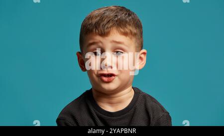 Mit weiten Augen und schockiertem Ausdruck scheint der Junge vor blauem Studiohintergrund überrascht oder neugierig zu sein, mitten im Gespräch, als ob er lustig erzählt Stockfoto