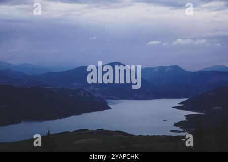 Panoramablick auf einen alpinen See, umgeben von Bergen in der Abenddämmerung, mit einem bewölkten Himmel und ruhigen Reflexen auf dem ruhigen Wasser. Stockfoto