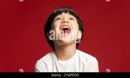 Kind, asiatischer Junge lacht mit zurückgeworfenem Kopf, weit geöffnetem Mund, voll im Moment eingetaucht vor kühlem rotem Studiohintergrund. Stockfoto