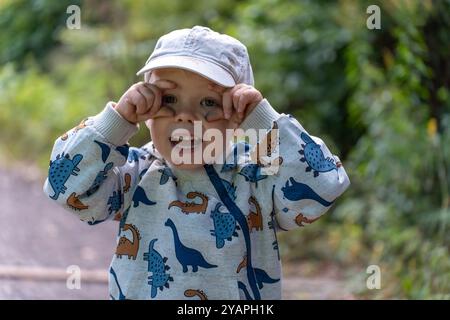 Niedliches Baby in Dinosaurierjacke und Hut, das an einem sonnigen Tag im Wald ein lustiges Gesicht macht. Stockfoto