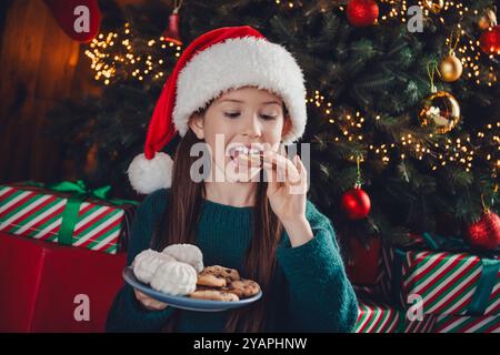 Foto eines netten kleinen Mädchens essen Snacks tragen grüne weihnachtskleidung genießen Sie gemütliche weihnachtsfeier Dekoration Innenraum Wohnung drinnen Stockfoto