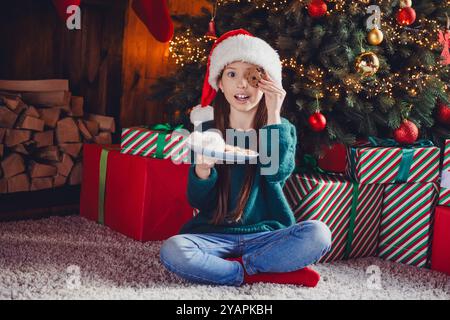 Ganzkörperfoto des kleinen Mädchens sitzen Boden essen Snacks tragen grüne weihnachtskleidung Genießen Sie die gemütliche Weihnachtsfeier Dekoration innen flach drinnen Stockfoto