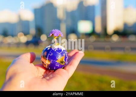 Die Hand hält ein kugelförmiges Puzzleteil, aus dem eine Blume wächst, vor einem verschwommenen urbanen Hintergrund, das das Wachstum der Umwelt symbolisiert. Stockfoto