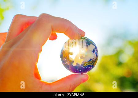 Die ausgestreckte Hand hält ein kugelförmiges Puzzleteil gegen Sonnenlicht und symbolisiert Einheit und Umweltbewusstsein in der Natur. Stockfoto