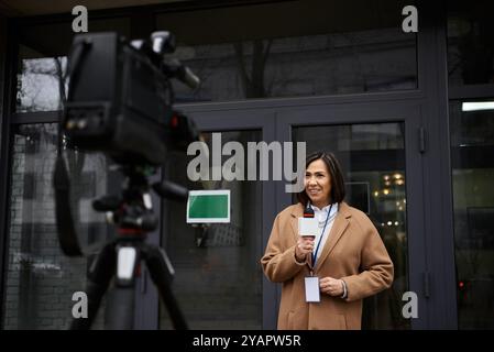 Eine multirassische Journalistin in einem beigefarbenen Mantel teilt aktiv Nachrichten, während sie draußen ein Mikrofon hält. Stockfoto