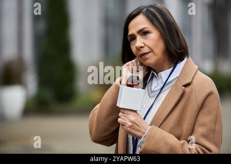 Eine multirassische Journalistin in einem beigefarbenen Mantel hält ein Mikrofon und berichtet mit Fokus draußen. Stockfoto