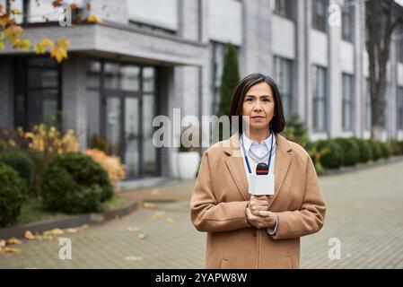 Eine multirassische Frau in einem beigefarbenen Mantel steht selbstbewusst mit einem Mikrofon und meldet sich vor Ort. Stockfoto