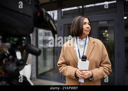 Ein multirassischer Journalist in einem beigefarbenen Mantel bereitet sich darauf vor, Nachrichten zu übermitteln, während er ein Mikrofon vor einem Gebäude hält. Stockfoto