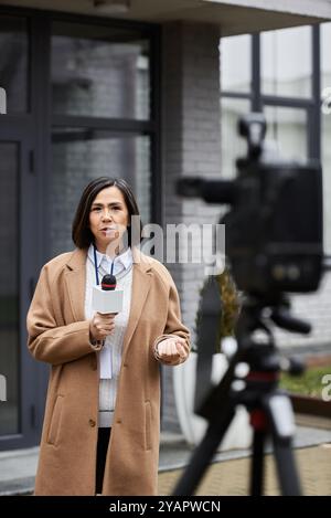 Der Journalist steht zuversichtlich, hält ein Mikrofon und berichtet außerhalb einer zeitgenössischen Struktur. Stockfoto