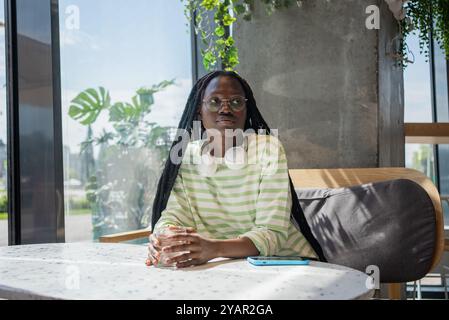 Die junge afroamerikanerin mit Kopfhörern sitzt allein an einem Tisch in einem Café, hält ein Glas Wasser und blickt in die Ferne Stockfoto
