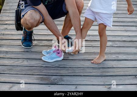 Vater hilft dem Kind, Socken und Schuhe anzuziehen, nachdem er barfuß auf einem Holzsteg gespielt hat. Die Beziehung zwischen Vater und Tochter Stockfoto