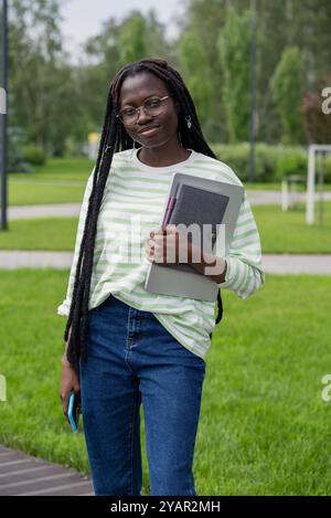 Junge afroamerikanische Frau, die selbstbewusst Technologie im Bildungsbereich einnimmt, mit Laptop, Notebook und Smartphone durch den Campus spaziert und für den Online-Unterricht in üppigem Grün gesäumt ist Stockfoto
