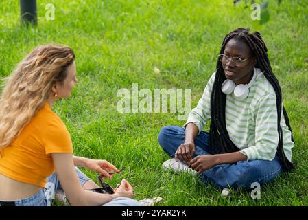 Zwei junge Frauen machen eine Pause vom Studium, plaudern auf Gras und genießen einen sonnigen Tag. In Gesprächen verwickelt, Geschichten geteilt, gelacht, entspannt draußen Stockfoto